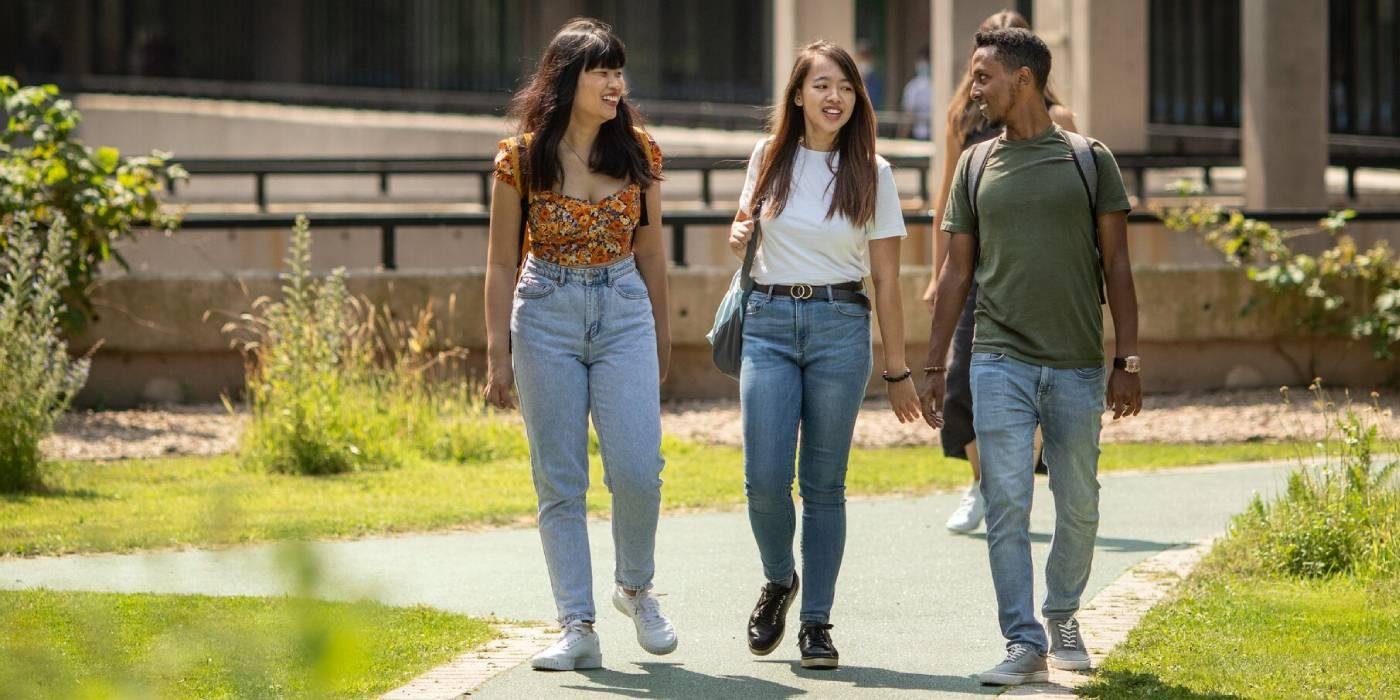 three students walk across the sunny campus and smile while talking.