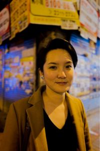 Photograph of Jamie Wong, taken at night/in the dark, pictured in front of a shuttered corner shop with bright signage above and on the shutters. Jamie Wong is smiling and wearing a beige-brown blazer, black top, a stud earring, shortish hair.