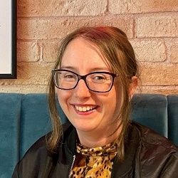 Profile photograph of Rebecca Porter, wearing a colourful yellow-black top, black jacket and black-framed glasses. She is smiling at the camera and is seated in front of an indoor light-brick wall and teal upholstery.