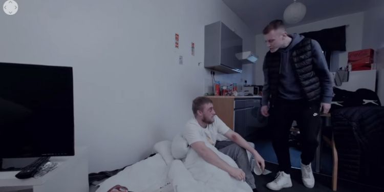 A man sits on a mattress on the floor. He has bruises on his inner arms and looks tired and scared. Another man looms over him as though he will shout at or hurt the man on the mattress.