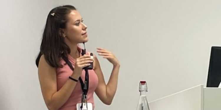 Woman in pink short sleeved top and black trousers speaks into microphone at the front of a lecture theatre.