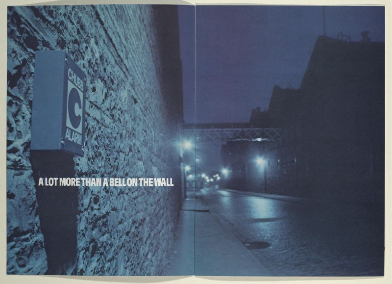 A double page magazine spread of a dark street in an industrial area with a Chubb alarm on a brick wall.
