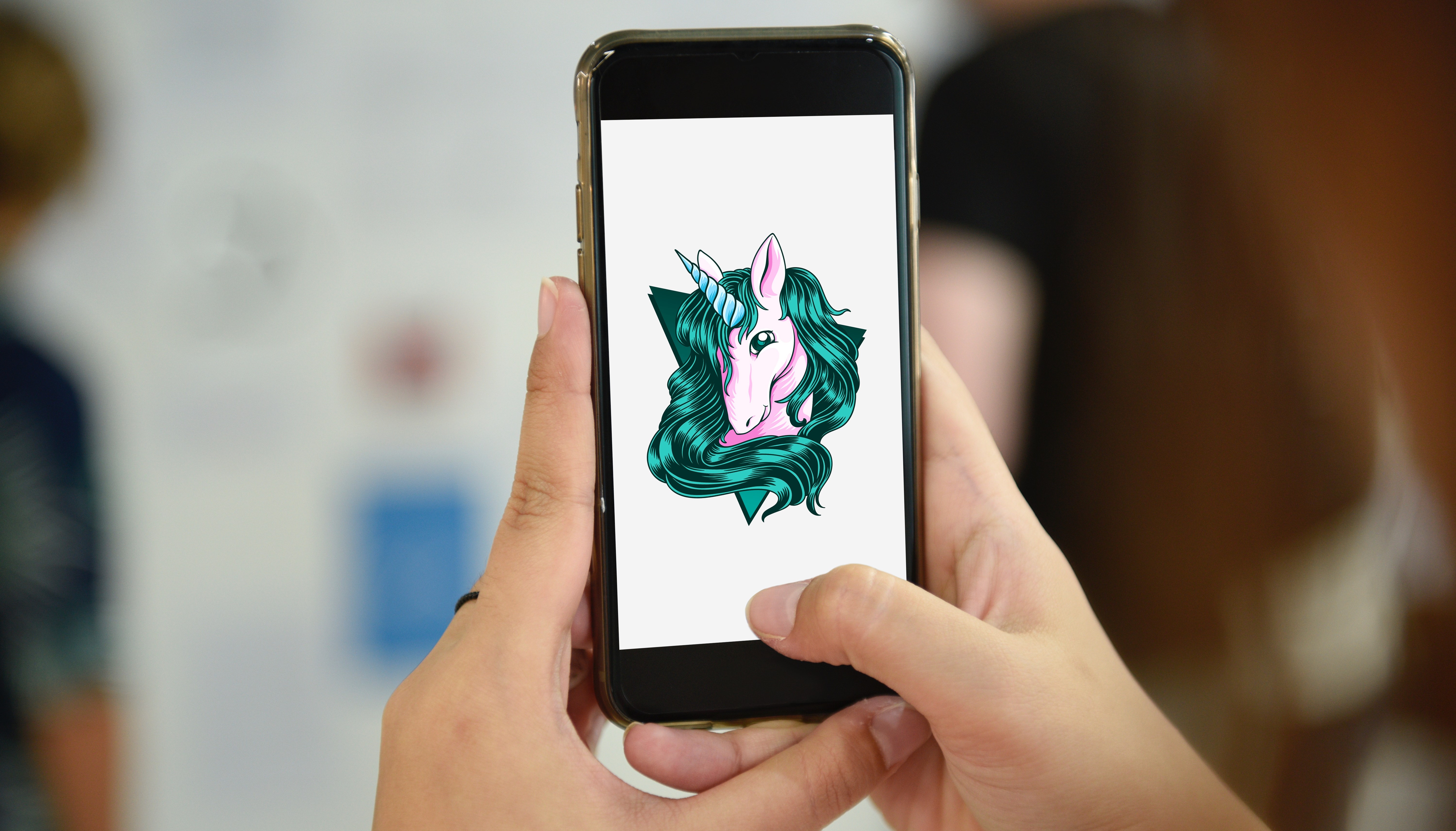 Photograph of two hands holding a phone with an illustration of a pastel pink unicorn's head with a green mane and pale blue horn, on a white background.