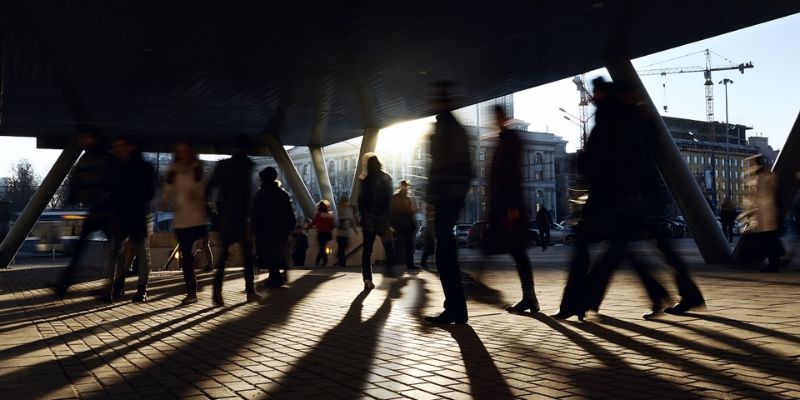 Approx. 20 people walking under a bridge, in an urban setting, each casting a long shadow towards the camera - figures are blurred as if walking quickly.