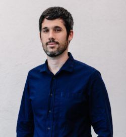 Profile photograph of Patricio Simonetto, standing against a plain background, looking at the camera, slightly smiling and wearing a beard and blue shirt.