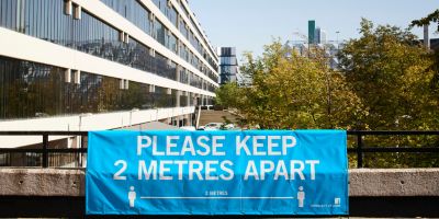 Outside shot of the University campus. A large blue banner is attached to a railing that reads: PLEASE KEEP 2 METRES APART.