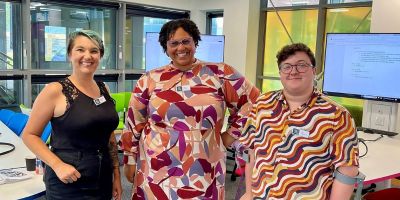 Three people in colourful smart-casual clothes stand together smiling broadly at the camera in a bright contemporary space with bold colours and lots of windows.