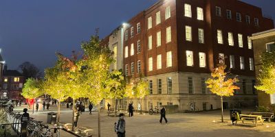 Union Square at the University of Leeds. People are walking through the square, there are trees lining either side of the square which are illuminated by fairy lights and Clothworkers' Building South is shown in the background