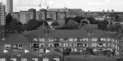 Black-and-white image of an urban housing estate with mid-rise residential buildings in the foreground and high-rise blocks, along with construction cranes, in the background.