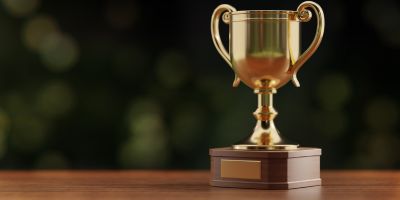 The image shows a gold trophy cup with ornate handles, sitting on a wooden base with a blank plaque. The background is dark with soft, out-of-focus lights.