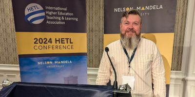 Nick standing at a podium with banners for the 2024 HETL Conference and Nelson Mandela University in the background. He is wearing a light-coloured shirt and lanyard, smiling toward the camera.