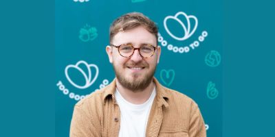 Jamie Crummie, a man with light brown hair and glasses, wearing a brown jacket over a white shirt, smiles at the camera. The background features a teal-colored wall with the 'Too Good To Go' logo and various eco-friendly symbols in white.