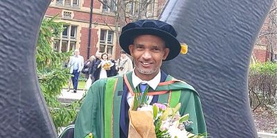 Dr Tesfalem Yemane stands in graduation cap and gown, holding a bouquet of flowers, in front of the sculpture in Beech Grove Plaza.