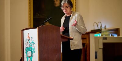 Woman in a black dress and grey cardigan stands at a podium delivering a lecture.