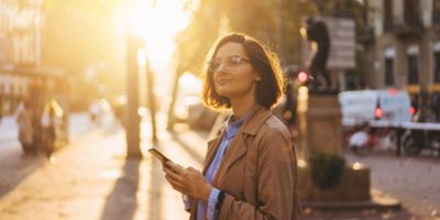 A woman with short hair, wearing glasses and a trench coat, stands on a city street bathed in warm, golden sunlight. She holds a phone in her hand, looking up with a calm and thoughtful expression. The background shows an urban setting.