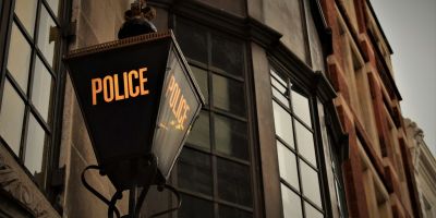 Traditional British police lantern mounted on a building, with the word 'POLICE' illuminated in yellow, set against the backdrop of a historic urban street with brick and glass architecture.