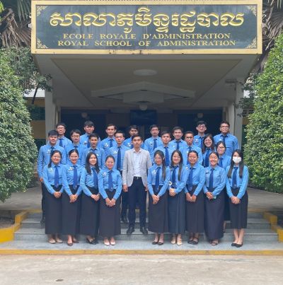 Bunthorn stands with his students below a sign that reads: 'Royal School of Administration'. He is in the middle surrounded by his students.