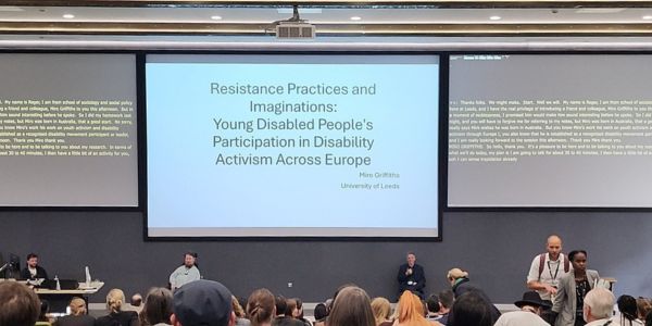 Photograph in auditorium, taken from audience seat, shows audience members sitting and panel members sitting/standing in front of Dr Miro Griffiths' keynote title slide. Live captioning slides are on either side.