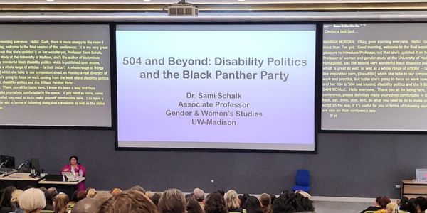 Dr Sami Schalk delivers her keynote. Title slide behind reads, "504 and Beyond: Disability Politics and the Black Panther Party". The auditorium seating is full.