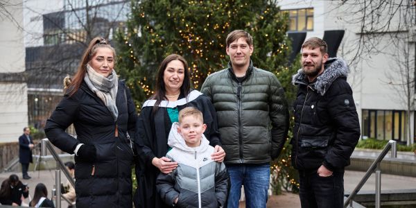 Sarah Lloyd in graduation gown stands with her family outdoors on University of Leeds Campus, the Beechgrove Plaza Christmas tree behind them.