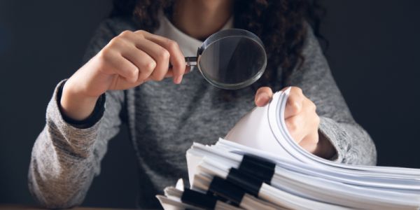 Woman is shown inspecting a pile of documents with a magnifying glass.