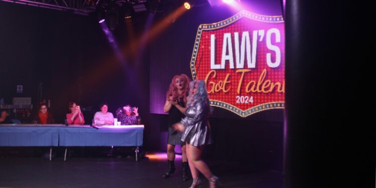 Two performers in vibrant costumes are on stage under bright lighting, engaging the audience during "LAW'S Got Talent 2024." The backdrop features the event's logo, and a panel of judges is seated at a table on the left, observing the performance.