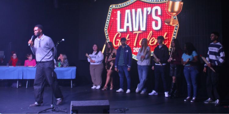 A host stands on stage with a microphone, speaking to the audience during "LAW'S Got Talent 2024." A group of participants stands in a row behind him, while the backdrop prominently displays the event's logo. A panel of judges is visible on the left side 