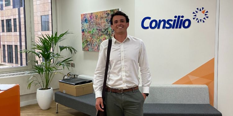 Carlo is standing in an office lobby with a bright smile. He is wearing a white dress shirt and grey pants, carrying a brown shoulder bag. Behind him is a colorful abstract painting and a sign on the wall that reads "Consilio." 