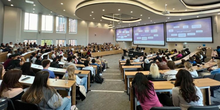A large lecture hall filled with students attentively listening to a presentation. The room has modern design elements, featuring large windows on the left, tiered seating, and three large projection screens displaying a slide titled "Why get involved?" 