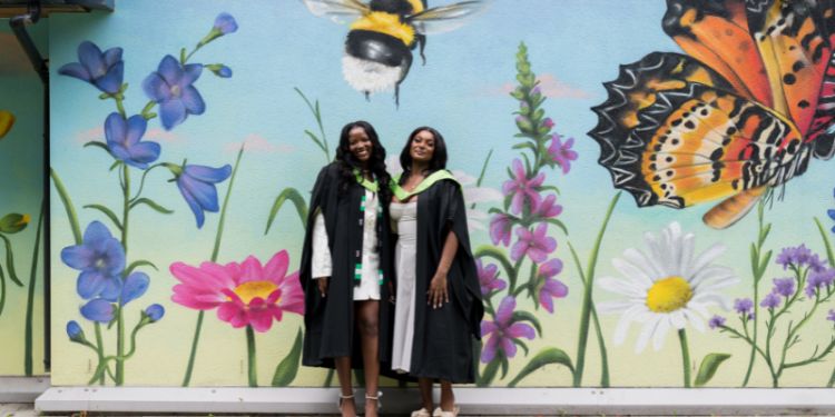 Two female graduates in gowns in front of mural wall painted with bees and butterflies