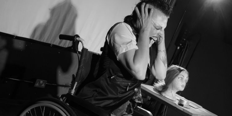 Drag artist Fae Fatale performs on stage. They are using a wheelchair and have their head in their hands, looking in pain. 