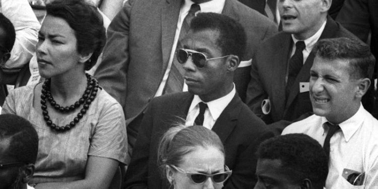 Photograph of James Baldwin, in suit and tie with sunglasses, sitting in an audience, watching something.