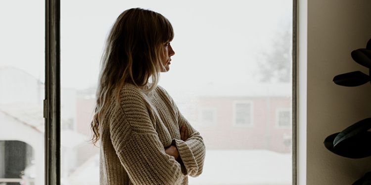 Woman looking sadly out of a window with her arms crossed. 