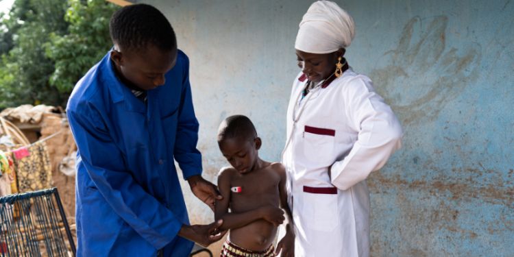 Medical trainee in blue workdress taking a small toddler's temperature under the close supervision of a pediatrician in a poor rural village in West Africa