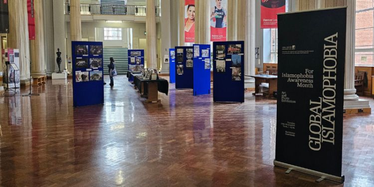 A wide-angled shot of Parkinson Court South shows multiple blue display boards with photographs, and tables in between with stand-alone framed photographs. In the foreground is a black exhibition banner with white text reading, "Global Islamophobia".