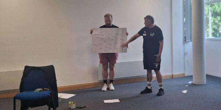 A staff member holds up a large piece of paper whilst a student gestures at it. Pens and paper are strewn throughout the room.