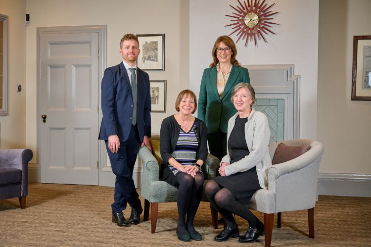 Four people, two seated and two standing. L-R standing: man in blue suit and woman in green suit. L-R seated: woman in striped dress and black cardigan, and woman in black dress and grey cardigan.