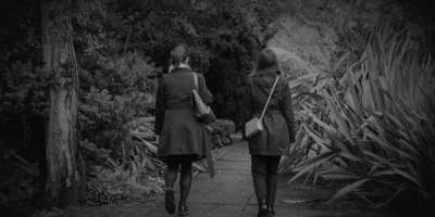 A greyscale photograph of two women walking in a park. We see them from behind.